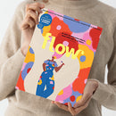 Person in hellbraunem Strickpullover hält die Zeitschrift „flow“ mit farbenfrohem Cover hoch; auf dem Titel steht „Das Magazin für persönliche Entwicklung, Kreativität & mehr Ruhe“ sowie „DIY-Girlande & Sommer-Journal“. Im Zentrum eine Illustration einer Person in blauer Latzhose mit bunten Farbklecksen, Pinsel in der Hand, vor abstrakt gemustertem Hintergrund in Rot, Gelb, Blau, Rosa und Beige.