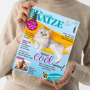 Person in beigefarbenem Pullover hält das Magazin „Geliebte Katze“, Ausgabe Juli 2026, vor neutralem Hintergrund. Titelbild zeigt eine silbergraue Katze in einer gelben Hängematte auf hellblauem Hintergrund. Lesbare Texte: „Einfach cool bleiben!“, „Katze Lini – Sie sollte nicht mehr leben“, „Tele-Medizin“, „Funktionale Snacks“, „Plötzlich Zoff!“, „Lieben wir unsere Katzen zu sehr?“ und „Großer Ratgeber Tatort Blumentopf“.