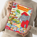 Person in beigem Pullover hält ein Kinderheft mit dem Titel „Benjamin Blümchen“. Auf dem farbenfrohen Cover ist der graue Elefant Benjamin in roter Kleidung und roter Mütze zu sehen, daneben Zeichnungen von Tieren und Pflanzen. Sichtbare Texte: „Gemüse-Snacks“, „So sehen Tiere“, „Doras Rechenpyramide“, „Ein fliegendes Chamäleon“. Logos: „Blue Ocean Entertainment“. Hintergrund in Gelb- und Grüntönen.