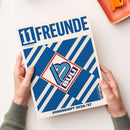 Blau-weiß gestaltetes Magazin „11FREUNDE“ mit dem Untertitel „Magazin für Fussballkultur“ und dem Aufdruck „SONDERHEFT 2026/27“, diagonal auf einem weißen Untergrund fotografiert. Zwei Hände mit grauen Strickärmeln halten das Magazin an den unteren Seiten; oben rechts liegt teilweise ein orangefarbener Gegenstand.