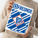 Eine Person ohne sichtbares Gesicht hält ein schräg abgebildetes Magazin mit weißem Rand und blau-weißen geometrischen Streifen. Auf dem Cover stehen „11FREUNDE“ und „Magazin für Fußballkultur“; mittig befindet sich ein rot umrandetes Quadrat mit einem blau-türkisen Logo und der Aufschrift „BULI“. Die Person trägt einen hellbeigen Strickpullover.