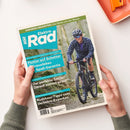 Magazintitel „ElektroRad“ in Weiß und Blau, gehalten von zwei Händen über hellem Hintergrund. Auf dem Cover eine person mit Helm und Sonnenbrille, fahrend auf einem schwarzen Gravelbike durch bewaldetes Gelände. Sichtbare Texte: „Flotter auf Schotter – Gravelbikes mit Spaß-Garantie“, „Der perfekte Sattel? Darauf kommt es an!“, „Rushhour-Tipps vom Verkehrs-Experten“ sowie Kopfzeile „Eurobike-Neuheiten: Das Beste von der Messe“.