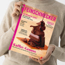 Person in beigem Strickpullover hält das Magazin „Feinschmecker“ mit pinkfarbenem Titelkopf vor neutralem Hintergrund. Das Cover zeigt gestapelte Stücke dunkler Schokolade vor goldenem Hintergrund, dazu Texte: „Schokolade! Die Sieger des großen Tafel-Tests“, „Trendziel Tokio“, „Südtirols Aufsteiger“, „Schnitzel & Schmankerl“ und „Kaffee-Genuss – Neue Maschinen und Geräte für jeden Geschmack“.