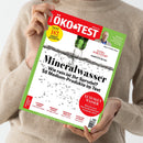 Person in beigem Strickpullover hält eine Ausgabe des Magazins „ÖKO-TEST“ mit rotem Titelbalken und grünem Rand. Hauptüberschrift: „Mineralwasser – Wie rein ist Ihr Sprudel? 56 Medium-Produkte im Test“. Weitere sichtbare Texte: „TEST 182 PRODUKTE FÜR JEDEN TAG“, „EXTRA SCHULSTART – KLEBESTIFTE IM TEST“, „LUXUSGUT WASSER – Warum auch Deutschland immer trockener wird“, Themen wie „ZITRONEN-SORBET“, „BARBECUE-Saucen“, „DEOSPRAYS OHNE ALU“, „AFTER-SUN-LOTIONEN“.