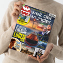 Person in beigefarbenem Strickpullover hält Magazin „Welt der Wunder“ mit blau-gelbem Titel. Auf dem Cover: großes Bild eines brennenden Öltankers, Bohrplattform, Männer in Anzügen, iranische Flagge und Diagramme. Hauptschriftzug: „Die größten Energielügen 2026“. Weitere Texte: „Der Sturm der Mongolen“, „Das wahre Facebook“, „Die kuriosen Verbrechen der Tiere“, gelbes Feld mit „Extra Mega-Poster – Die Inseln unserer Welt“.