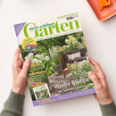 Titelbild der Gartenzeitschrift „mein schöner Garten“, Ausgabe Juli 2026. Im Zentrum steht ein sommerlicher Garten mit weißen Blüten, Sitzbank mit grauen Kissen und Holztisch auf Steinboden. Textelemente: „Weiße Blüten im Sommergarten“, „Hitzefeste Sukkulenten“, „Alles Wichtige in diesem Monat“ sowie Themen zu Gartenduschen, Brunnen, Obstsorten und Sommersträuchern. Hände halten das Magazin über einer hellen Fläche.