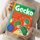 Person in beigem Strickpullover hält ein Magazin mit grün-orangefarbenem Titelblatt „Gecko – Die Bilderbuchzeitschrift“ (Nr. 114). Sichtbare Texte: „PIZZA Sechs Jahreszeiten“, „JUPP und die Gemüsegäng“, „BROKKOLI im All“, „Geschichten, Sprachspiele und Mitmachseiten“, „Alle zwei Monate neu ohne Werbung“, „THEMA: Gemüse!“. Illustration mit drei Brokkoli-Figuren in Astronautenanzügen und kleinen Raketen auf orangefarbenem Hintergrund.