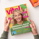 Titelbild der Zeitschrift „vital“ (April/Mai 2024) mit großem Porträt einer lächelnden Frau mit blondem Haar und grünem Pullover vor gelbem Hintergrund. Aufschriften u. a.: „Abnehmen durch besseren Schlaf“, „So schmeckt Spargel am besten“, „Sonnenschutz für jeden Hauttyp“, „Wie die Schulter wieder rund läuft“, „Heilsame Orte“ und „Schon wieder Sonnenbrand?“. Hände halten das Magazin auf hellem Tisch mit orangefarbenem Tablett oben rechts.