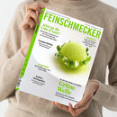 Helle Zeitschrift „FEINSCHMECKER“ mit grün-weißer Titelseite, gehalten von einer Person in beigem Wollpullover vor neutralem Hintergrund; mittig ein rundes, hellgrün überzogenes Dessert mit Kräuterdekoration auf weißem Teller, Texte u. a.: „Jetzt an die Côte d’Azur!“, „Sommerküche“, „Grüne Welle – 30 Seiten über Olivenöl“, „Hamburg – Koch des Monats“ und „Kaffee-Metropolen“.