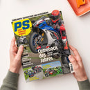 Titelblatt des Magazins „PS sport motorrad magazin“ mit großem Foto eines Fahrers auf einer Suzuki GSX-R 1000 R in Blau, Weiß und Rot beim Wheelie. Schlagzeilen: „Comeback des Jahres“, „Vergleich Superbikes“, „Vergleich Naked Bikes“ sowie Hinweise zu Modellen Yamaha R1, Bimota KB4 RC und Metzeler Sportec O1 RS. Gehalten von zwei Händen über hellem Tisch mit orangefarbenem Tablett.
