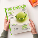 Titelbild des Magazins „Feinschmecker“, gehalten von zwei Händen auf hellem Untergrund. Dominierende Farben Weiß und Grün. Zentrale Abbildung: rundes, grün bepudertes Dessert mit Kräuterdekoration. Sichtbare Texte: „Jetzt an die Côte d’Azur!“, „Sommerküche“, „Grüne Welle“, „Hamburg“, „Gardasee“, „Kaffee-Metropolen“. Magazinrand mit grünem Logo und senkrechtem Schriftzug „FEINSCHMECKER“.