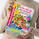 Hellhäutige Person in beigefarbenem Strickpullover hält eine Ausgabe der Kochzeitschrift „kochen & genießen“ mit rotem Seitenbalken. Titelthemen: „Turbo-Hits für den Sommer“, „Dinner im Garten mit Pute, Pasta & Party-Spritz“, „So handlich! Mitbringkuchen“. Deckblatt zeigt einen Teller mit gegrilltem Fleisch, buntem Salat, Holztisch im Hintergrund und blaue Hortensien als Dekoration.