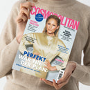 Frau in beigefarbenem Pullover hält ein Exemplar der Zeitschrift „Cosmopolitan – The German Issue“ mit rosa Titelaufschrift. Auf dem Cover eine blonde Frau in glänzender, cremefarbener Bluse mit verschränkten Armen. Sichtbare Texte: „PERFEKT WAR NIE DER PLAN“, „Selbstsabotage erkennen, verstehen“, „WILDES NEID-LIFE“, „POOL, PASSION, PANIK“ sowie weitere Schlagzeilen zu Mode, Reisen und Wellness auf hellblauem Hintergrund.