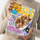 Person in beigefarbenem Strickpullover hält ein Magazin mit dem Titel „essen & trinken“. Das Cover zeigt einen weißen Teller mit mediterranem Gericht aus Fleischspießen, roten Zwiebeln, Kräutern und Paprika auf blau-weißer Tischdecke. Sichtbare Texte: „Mediterrane Urlaubsküche“, „Mozzarella, Burrata & Co.“, „Himbeerträume“, „Saucen-Schule“, „Die Pfalz für Genießer“.