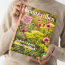 Titelblatt der Zeitschrift „kraut&rüben“ Ausgabe 07/2026, gehalten von einer Person in beigefarbenem Pullover vor neutralem Hintergrund. Cover zeigt eine Nahaufnahme gelber und pinker Sommerblumen auf einer Wiese, mit Texten wie „Tomatenliebe – Pflege-Leitfaden für köstliche Früchte“, „Griechisches Sein“, „Zukunftsspeicher“ und Haupttitel „Jeder Tag ein Sommerfest!“.