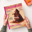 Titelbild der Zeitschrift „Feinschmecker“, gehalten von zwei Händen über einer hellen Oberfläche; Cover zeigt gestapelte Stücke dunkler Schokolade, mit herabfließender flüssiger Schokolade, vor cremig-beigem Hintergrund. Sichtbare Texte: „SCHOKOLADE!“, „Kaffee-Genuss“, „Trendziel Tokio“, „Südtirols Aufsteiger“, „Schnitzel & Schmankerl“ sowie Logo und Ausgabeangaben am oberen Rand.