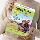 Person in beigefarbenem Strickpullover hält das Magazin „Spotlight – Das Magazin der englischsprachigen Welt“ in beiden Händen. Titelcover mit grünem Kopfbereich zeigt ein lachendes Paar, die Person vorne trägt Sonnenbrille und Blumenkranz, im Hintergrund Festivalwiese mit Menschen und britischer Flagge. Textaussagen u. a.: „CELEBRATING THE SUMMER 2026“, „The 15 best events in the English-speaking world“, „UNDERSTANDING THE USA“, „A DAY IN INVERNESS“, „THE MADNESS OF CRICKET“.
