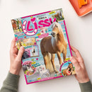 Titelseite der Zeitschrift „Lissy“ mit pinkem Rand und Herzsymbolen, im Mittelpunkt ein braunes Pferd mit heller Mähne am Strand. Rundherum kleinere Bilder von Pferden, einer Frau mit Pferd sowie Zeichnungen und pink-gelbe Textflächen mit Aufschriften wie „Retro Comic“, „4 Poster zum Verlieben“, „Rezept für deine Einhorn-Party“ und „Wissen Pferde ♥ Bienen“. Zwei Hände halten das Magazin über hellem Tisch.