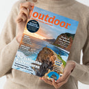 Zeitschrift „outdoor – Wandern. Reisen. Abenteuer.“ wird von einer Person in beigefarbenem Pullover vor neutralem Hintergrund gehalten; Titelbild zeigt eine felsige Küstenlandschaft mit Meer und Sonnenuntergang. Sichtbare Texte: „Die schönsten Küstentouren – Traumwanderungen von Norwegen bis Kroatien“, „Peaks of the Balkans“, „WILDES TRENTINO“, „ZELTE IM STURM“ und ein blauer Kreis mit „Testsieger! Die besten Produkte 2026“.