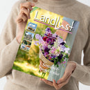Person in beigem Strickpullover hält eine Ausgabe der Zeitschrift „LandLust“ mit floraler Titelseite. Das Cover zeigt einen bunten Blumenstrauß aus violetten, rosa, weißen und roten Blüten vor grün-gelbem Hintergrund, dazu Texte wie „Sommer-Geschenke“ und Themen zu Kräutern, Kuchenrezepten und Gartenbau. Am linken Rand kleine Fotos mit Landschaft, Gebäude, Gartenmotiv und Schriftzug „LEVI“.