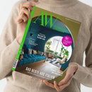 Eine beige gekleidete Person hält eine Ausgabe des Magazins „AW Architektur & Wohnen“ mit grün-gelbem Cover, großem grünen „AW“-Logo und pinkem Kreis mit der Aufschrift „AW Architekt des Jahres 2026“. Abgebildet ist ein modern eingerichtetes Wohnzimmer mit blauer Couch, Holzboden, runder Wandöffnung und Pflanzen. Texte: „Das Beste aus Italien“, „Unterwegs Mailand für Design-Fans“, „Dossier: Erlebt der Plattenbau einen Imagewandel?“