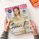 Titelblatt der Zeitschrift „Cosmopolitan“ mit einer blonden Frau in glänzender beiger Bluse vor hellgrauem Hintergrund, gehalten von zwei Händen. Aufschriften u. a.: „THE GERMAN ISSUE“, „PERFEKT WAR NIE DER PLAN“, „POOL, PASSION, PANIK“, „WILDES NEID-LIFE“, „Wellness oder Wahnsinn?“ sowie Themenbereiche zu Mode, Urlaub, Fitness und Lifestyle in farbigen Schriftfeldern in Pink, Blau und Schwarz.