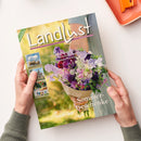 Titelblatt der Zeitschrift „Landlust“, gehalten von zwei Händen über einer hellen Tischfläche; im Mittelpunkt ein Strauß aus violetten, rosafarbenen und weißen Blumen, darüber die Aufschriften „Selbst gebaut: Pflanzkasten für Kräuter“, „Fruchtig: Kuchen ohne Backen“, „Rettich: Anbauen und ernten“ sowie unten „Sommer-Geschenke“, links kleinere Bilder mit Kuh, Gebäude und Gartenmotiv.