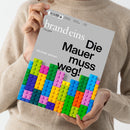 Person in beigem Strickpullover hält das Magazin „brand eins – Das Geld“ mit grauem Titelblatt, schwarzer Überschrift „Die Mauer muss weg!“, Unterzeile „Modular denken“ und einer grafischen Darstellung aus bunten, übereinandergestapelten Legosteinen in Grün, Blau, Gelb, Orange, Rosa, Lila und Türkis vor neutralem Hintergrund.
