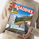 Helle, beige gekleidete Person hält das Magazin „ROADBIKE“ mit roter Titelzeile vor neutralem Hintergrund; auf dem Cover fahren vier Radsportler auf einer Bergstraße bei sonnigem Wetter, umgeben von grünen Hängen und blauem Himmel. Sichtbare Texte: „LEICHT & SCHNELL“, „FITNESS“, „Elektro-Pumpen“, „Sport-BHs“, „SCHÖNES ITALIEN“, „GUT & GÜNSTIG“, sowie verschiedene Testankündigungen zu Rennrädern und Zubehör.