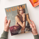 Titelblatt der Zeitschrift „MADAME“ mit orangefarbener Überschrift; im Zentrum sitzt eine Frau mit langem, hellbraunem Haar in dunkelroter Spitzenkleidung auf einem Holzstuhl. Text auf dem Cover: „SPECIAL REISE LUST“, „Japans schönste Routen“, „Dänemark – der Insider“, „Kopenhagen, Europas Stilmetropole“ sowie „BEACH-WEAR & ACCESSOIRES MIT OCEAN FLAIR“. Hände in grünem Pullover halten das Magazin über weißem Hintergrund.