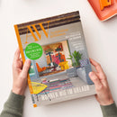 Titelbild der Zeitschrift „AW Architektur & Wohnen“ mit großem orangefarbenem „AW“-Logo oben, grauer Hintergrundfläche und Foto eines modern möblierten Wohnraums mit farbenfrohem Wandbild, gelbem und orangefarbenem Stuhl, Pflanze, Teppich und Couch. Aufdrucke u. a.: „52 Seiten Bad & Wellness“, „Neue Architektur aus Dänemark“, „Kunstvolle Muster & Texturen“ und „Wohnen wie im Urlaub“. Zwei Hände halten das Heft.