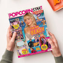 Titelblatt des Magazins „Popcorn Star & Poster“ mit dem Hauptmotiv einer jungen Frau mit blonden Haaren vor türkisblauem Strand- und Wasserhintergrund. Farbige Schriftzüge: „Fun am Beach – wo Stars den Sommer feiern!“, „16 coole Poster“, „Mega-Hype um BTS“, „Spidey is back!“ sowie „2 Mega Poster Lifesize!“. Mehrere kleinere Fotos von Prominenten und Magazininhalten sind rund um das Hauptbild platziert. Zwei Hände halten das Heft über hellem Untergrund.