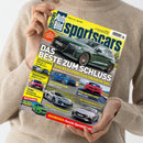 Person mit beigefarbenem Strickpullover hält das Magazin „Auto Bild sportscars“ mit gelbem Titelhintergrund und blauer Schrift. Auf dem Cover sind mehrere Sportwagen abgebildet, großformatig ein grüner Audi RS 3 mit der Überschrift „Das Beste zum Schluss – Audi RS 3 competition limited im Test“ sowie kleinere Bilder von Fahrzeugen wie Corvette, Renault, BMW und Ford. Weitere sichtbare Texte: „Hausbesuch: Porsche-Spezialisten“, „Technik“, „Neue Rubrik“.