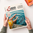 Zwei Hände in grünen Strickärmeln halten die Zeitschrift „Servus – einfach gut leben“. Auf dem Titelbild sind Beine einer weiblich gelesenen Person zu sehen, die auf einem hölzernen Steg sitzt und die Füße ins Wasser taucht. Haupttitel: „Warum Wasser Wunder wirkt – Gesunde Rituale und quellfrische Erkenntnisse“, daneben roter Kreis mit Text „So schmeckt die Kindheit“, hellblauer Hintergrund mit Wellenstruktur.