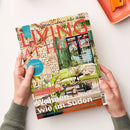 Magazintitel „LIVING AT HOME“ in großer roter Schrift auf einem Cover mit mediterraner Steinhausfassade, grünen Fensterläden und einer Terrasse mit grüner Couch und Dekorationen. Weitere sichtbare Texte: „Wohnen wie im Süden“, „Citytrip München“, „Sommerliche Ideen für den Tisch, Geschirr, Stoffe, Accessoires & DIY“, „Urlaubsfrische“, „Mein Zuhause voller Ideen“. Zwei Hände halten das Magazin über hellem Untergrund.