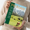 Eine hellhäutige, langärmlig bekleidete Person hält eine Zeitschrift mit grünem und hellblauem Cover; Aufschrift „MERIAN – The Art of Travel“ und Haupttitel „Sommer in den Alpen“. Sichtbar sind eine grüne Almwiese mit einer braun-weißen Kuh, verstreute Hütten, ein Waldstreifen und ein felsiges Bergmassiv im Hintergrund. Linke Coverseite mit Texten zu „Mexico City“, „Korfu“, „München“ und „Teneriffa“.