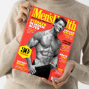 Person in beigefarbenem Strickpullover hält ein Magazin mit rotem Hintergrund und gelbem Titel „Men’s Health – Die große Jubiläums-Ausgabe“. Auf dem Cover ein muskulöser, oberkörperfreier Mann in Schwarz-Weiß Aufnahme. Textausschnitte: „Stärker in jedem Alter“, „Endlich schlank“, „Super? Mann!“, „Härte-Test“, gelbes Sechseck mit schwarzer Aufschrift „30 Jahre Men’s Health“, weiße und gelbe Überschriften auf rotem Grund.