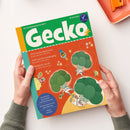 Titelblatt der Kinderzeitschrift „Gecko“ Nr. 114 mit grünem Kopfbereich und rotem Hintergrund, darauf gezeichnete Brokkoli-Figuren in Astronautenanzügen, eine fliegende Rakete und kleine Planeten. Sichtbare Texte: „PIZZA Sechs Jahreszeiten“, „JUPP und die Gemüsegang“, „BROKKOLI im All“, „Thema: Gemüse!“. Gehalten von zwei Händen vor hellem Untergrund, rechts orangefarbene Schale mit Büroklammern.