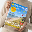 Person in beigem Strickpullover hält das „ADAC Reisemagazin“ mit blau-gelbem Logo. Das Cover zeigt einen Sandstrand mit Dünen, einer dänischen Flagge und einem kleinen Haus unter blauem Himmel. Sichtbare Texte: „Sommerglück in Dänemark“, „COOLCATION URLAUB OHNE HITZE“, „Kärnten neu erleben“, „Traumreise: Kanada“ und „Auf nach Paris!“. Farbtöne dominieren in Blau, Gelb und Weiß.