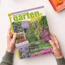Titelblatt der Gartenzeitschrift „gartenspaß“ Juni 2025, gehalten von zwei Händen über einer hellen Tischfläche; Hauptmotiv zeigt einen blühenden Garten mit violetten Hortensien, Weg und Rankbogen; sichtbare Texte u. a. „Neue Sorten für jeden Standort – Zauberhafte Hortensien“, „Gärtnern leicht gemacht“, „Gestalten mit Rank- & Rosenbogen“ sowie Themen zu Gartengestaltung, Bienemagneten und Pflanztipps.