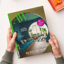Magazin „AW Architektur & Wohnen“ mit olivgrünem Titelcover und großem kreisförmigem Innenarchitektur-Foto: im Vordergrund ein dunkelblaues Sofa mit Kissen, im Hintergrund ein heller Wohnraum mit grüner Couch, Holzstühlen, Teppich und großen Pflanzen. Sichtbare Aufschriften: „Unterwegs Mailand für Design-Fans“, „Das Beste aus Italien“, „AW Architekt des Jahres 2026“, „Wilde Schönheit – Stauden, Struktur und Sinnlichkeit“.