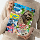 Beige gekleidete Person hält ein Magazin mit dem blauen Titel „Galileo Genial“. Auf dem Cover ist ein Faultier in Nahaufnahme vor grünem Hintergrund zu sehen, rechts daneben der Schriftzug „Tierisch faul oder nur entspannt?“. Weitere Bilder zeigen einen bunten Fidget-Skorpion, eine Person im Schnee mit der Überschrift „Gletscher in Not“ sowie kleinere Fotos von sportlichen und medizinischen Themen.