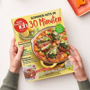 Titelblatt der Kochzeitschrift „meine Familie & ich“ mit gelbem Hintergrund, großem Schriftzug „SOMMER-HITS IN 30 Minuten“ und Foto eines Gerichts aus Hähnchen, Gnocchi, Tomatensoße und Kräuterpesto in runder Schale. Textaufdrucke u. a. „EXTRA-HEFT Preiswert kochen“, „Familienkuchen“, „Mit Beeren & Obst“. Gehalten von zwei Händen mit grünem Strickpulli über hellem Tisch, orangefarbenes Tablett im Hintergrund.
