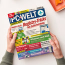 Magazintitel „PC-WELT+“ in Blau-Weiß mit Zusatztexten in Rot, Gelb, Grün und Weiß auf mehrfarbigen Hintergrundfeldern; zentrale Schlagzeile „Registry-Tricks mit Sofortwirkung“, weitere Themen „Windows nach Maß“, „Soforthilfe-Paket“, „Anti-Phishing-DVD“ und „Pannenhilfe Windows 11“; zwei menschliche Hände halten das Heft auf hellem Tisch neben orangefarbenem Behälter.