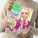 Person in beigefarbenem Strickpullover hält das Magazin „TOPModel“ mit pastellfarbenem Cover. Darauf gezeichnete weibliche Figur mit langen blonden Haaren, violettem Pullover und Herzhandgeste. Sichtbare Texte: „Knusper Frühstück“, „Ein unvergesslicher Geburtstag“, „Happy Ostern“, „Stylecheck“, „Deine kreative Lieblings-Welt“, „TOPModel by Depesche“, „Deine Extras! Hasen-Schachtel“, „Sammelposter“.