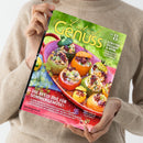 Person in beigefarbenem Strickpullover hält das Magazin „Lust auf Genuss“ Ausgabe Nr. 8 in den Händen; Titel zeigt bunt gefüllte Tomaten und runde Zucchini auf rotem Teller vor grünem Hintergrund, mit den Texten „Tomaten, Zucchini & mehr – Köstlich-kreativ kombiniert mit Fleisch & Fisch“ und „Die beste Zeit für Sommergemüse!“.