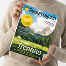 Helle Zeitschrift mit dem Titel „ADAC Reisemagazin“ in schwarzen Buchstaben auf blauem Hintergrund, gehalten von einer Person in beigefarbenem Strickpullover. Cover mit Berg- und Waldlandschaft, darüber Texte u. a.: „Lust auf Italien! Trentino“, „Kroatiens Inseln“, „Schweden erleben“, „City-Guide: Prag“ und gelber Kreis mit Hinweis „14 Seiten Extra Abenteuer-Reisen“.
