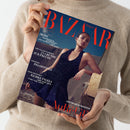 Magazin „Harper’s Bazaar“ mit rotem Schriftzug „BAZAAR“ im oberen Bereich, gehalten von einer Person in beigefarbenem Strickpullover. Auf dem Cover eine feminine Person in dunklem, glitzerndem Kleid vor Felsenlandschaft. Sichtbare Texte: „MODE – SONNENGELB, SEEROSE, SHORT UND SCHMUCK ZUM VERLIEBEN“, „DER KULTUR-SOMMER – 23 GRÜNDE ZUR FREUDE“, „PARADIES – PELOPONES“, „BJARKE INGELS – FORM VON LUXUS“, sowie „GNTM-GEWINNERIN“.