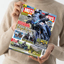 Eine Person in beigem Strickpullover hält ein Magazin mit dem Titel „MOTORRAD NEWS“ in beiden Händen, Titelthema „Frau Strom ist zurück“. Auf dem Cover ein großes Foto eines Fahrers oder einer Fahrerin auf einer blauen Suzuki SV-7GX, umgeben von kleineren Bildern weiterer Motorräder, Landschaftsszenen und Zubehör. Sichtbare Texte: „FAHRBERICHT SUZUKI SV-7GX“, „KAUFHILFE“, „SZENE REPORTAGEN“, „100 Jahre Ducati“, „IST TEURER BESSER?“ sowie diverse Modellnamen.