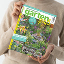 Helle Person in beigem Strickpullover hält die Juli-2026-Ausgabe des Magazins „Gartenspaß“ mit grün-gelbem Titel. Das Cover zeigt einen bunt blühenden Cottage-Garten mit Holztisch und Stühlen, umgeben von violetten, gelben und weißen Blumen. Textelemente u. a.: „Blütenfest im Cottage-Garten“, „Tolle Taglilien“, „Schritt für Schritt“, „Süße Sommerfrüchte“ und „Schatten­glück charmante Pflanzideen“.